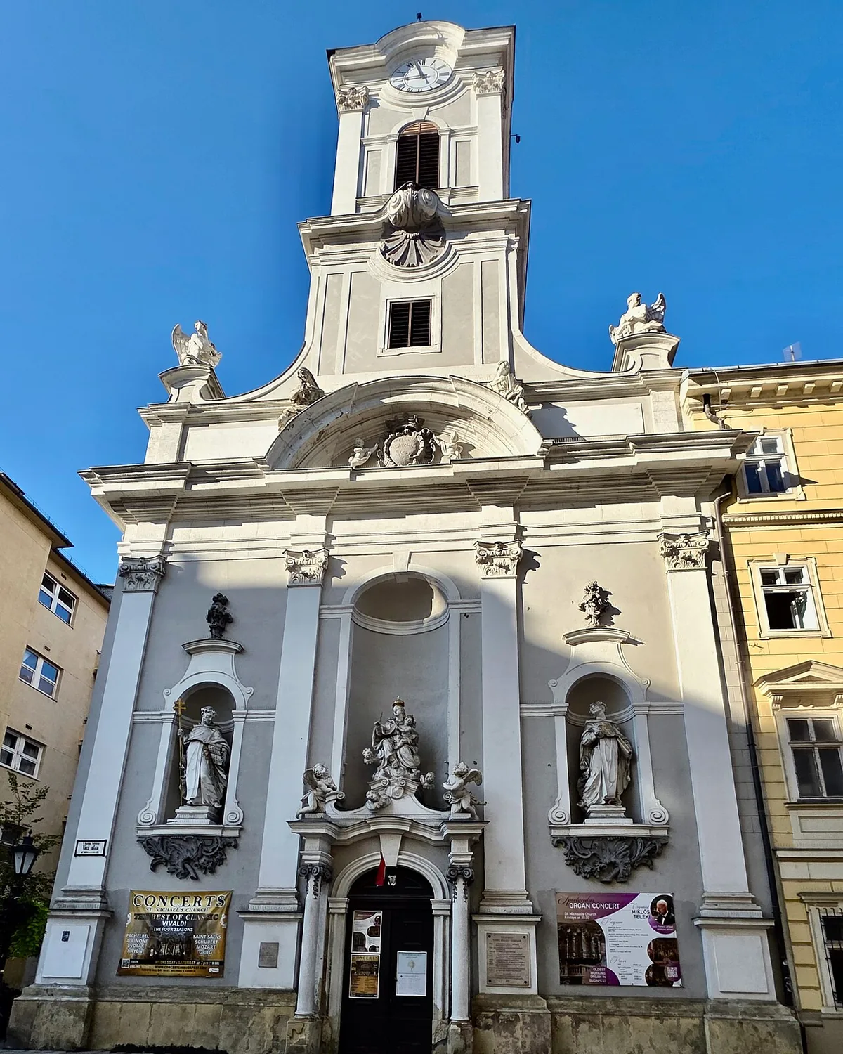 Kirkens hvide barokfacade på Váci utca i Budapest, med et klokketårn, statuer i nicher og et par søjler ved døråbningen under en blå himmel