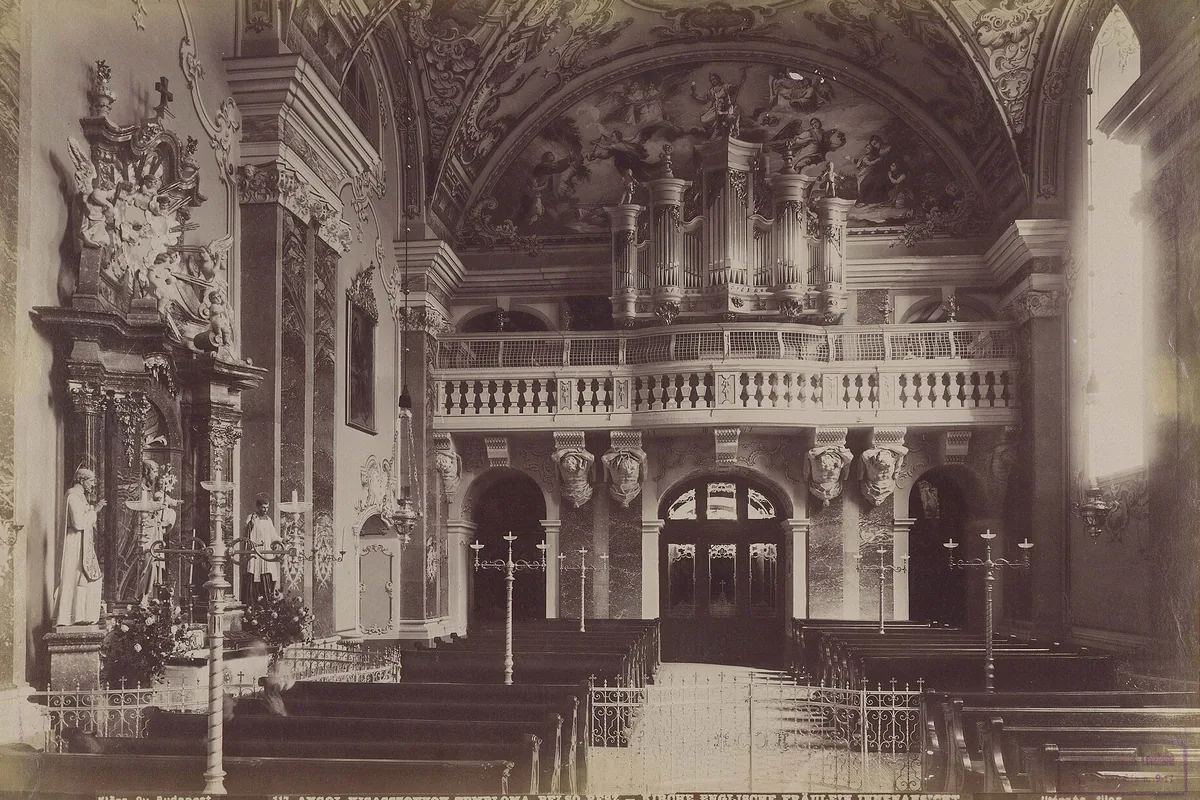 Sepiová fotografie interiéru kostela kolem roku 1892 s varhanní galerií nad balustrádou, oltářem vlevo a řadami lavic vpředu