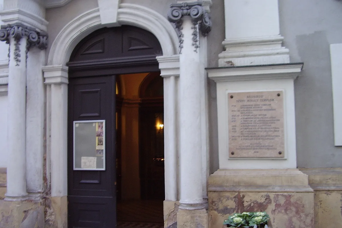 Klenutý kamenný portál kostela na budapešťské ulici, po obou stranách sloupy, nástěnka a kamenná pamětní deska