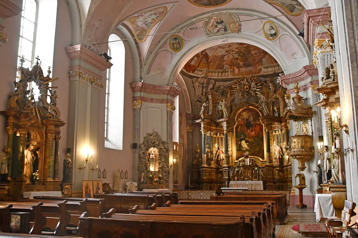 Loď barokního kostela v Budapešti s řadami tmavých dřevěných lavic obrácených k pozlacenému hlavnímu oltáři pod růžově vymalovanými klenbami s freskami