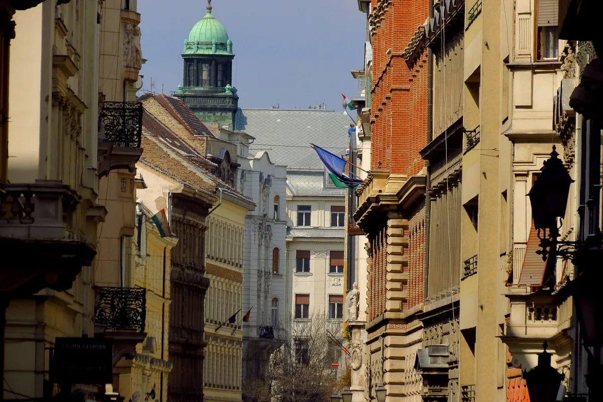 Váci utca curving between tall buildings, with a green copper dome and flags in the distance