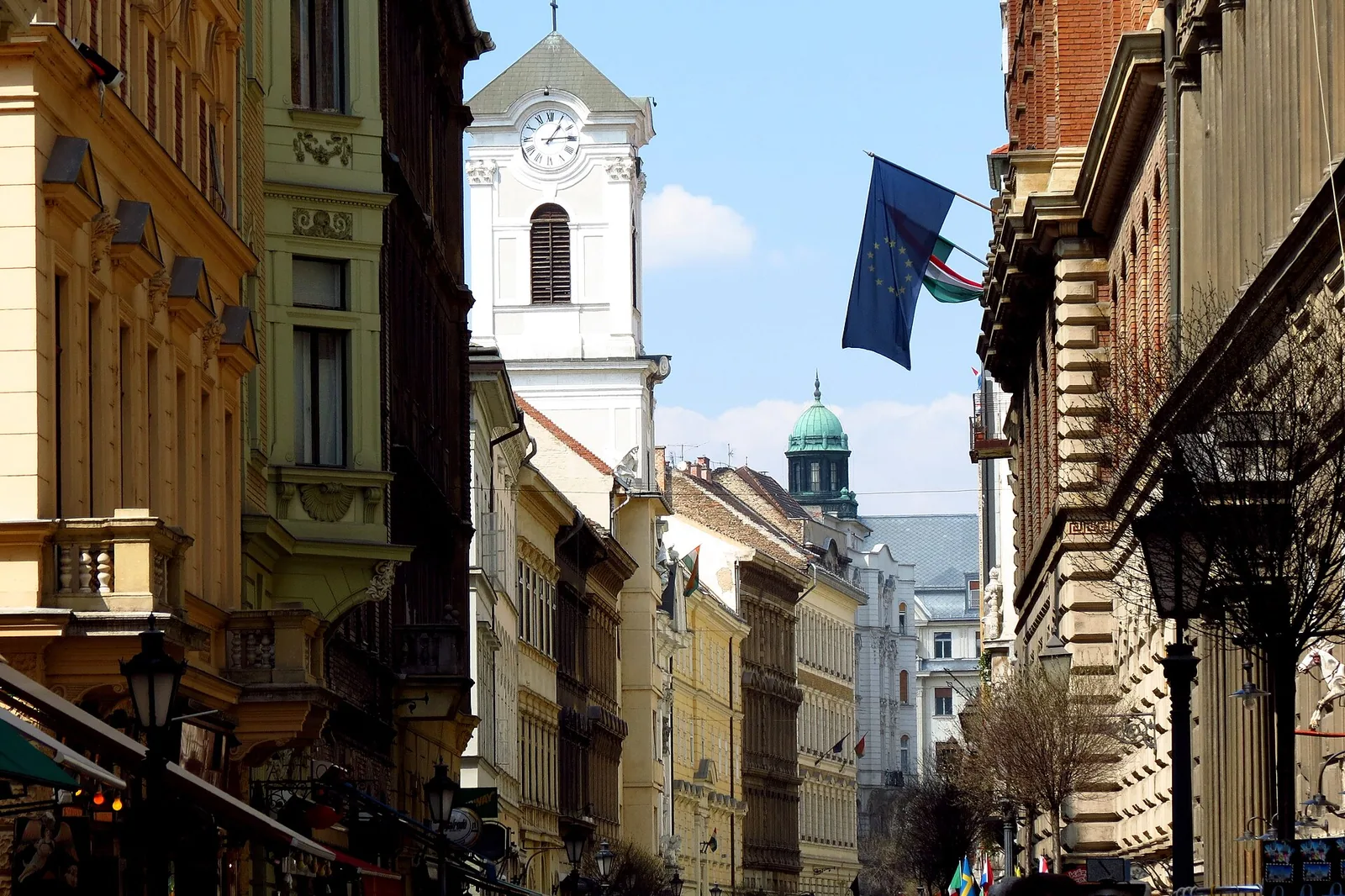 Váci utca set mod kirken, en smal gågade mellem høje bygninger fra det nittende århundrede, med det hvide klokketårn, der rejser sig forude, og et EU-flag over gaden